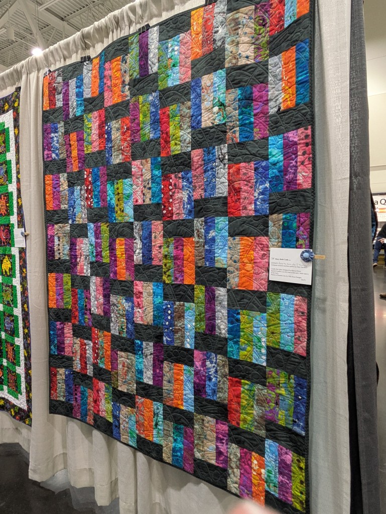 A colorful patchwork quilt displayed at an exhibition, featuring various rectangular fabric pieces in vibrant shades, surrounded by a dark border.