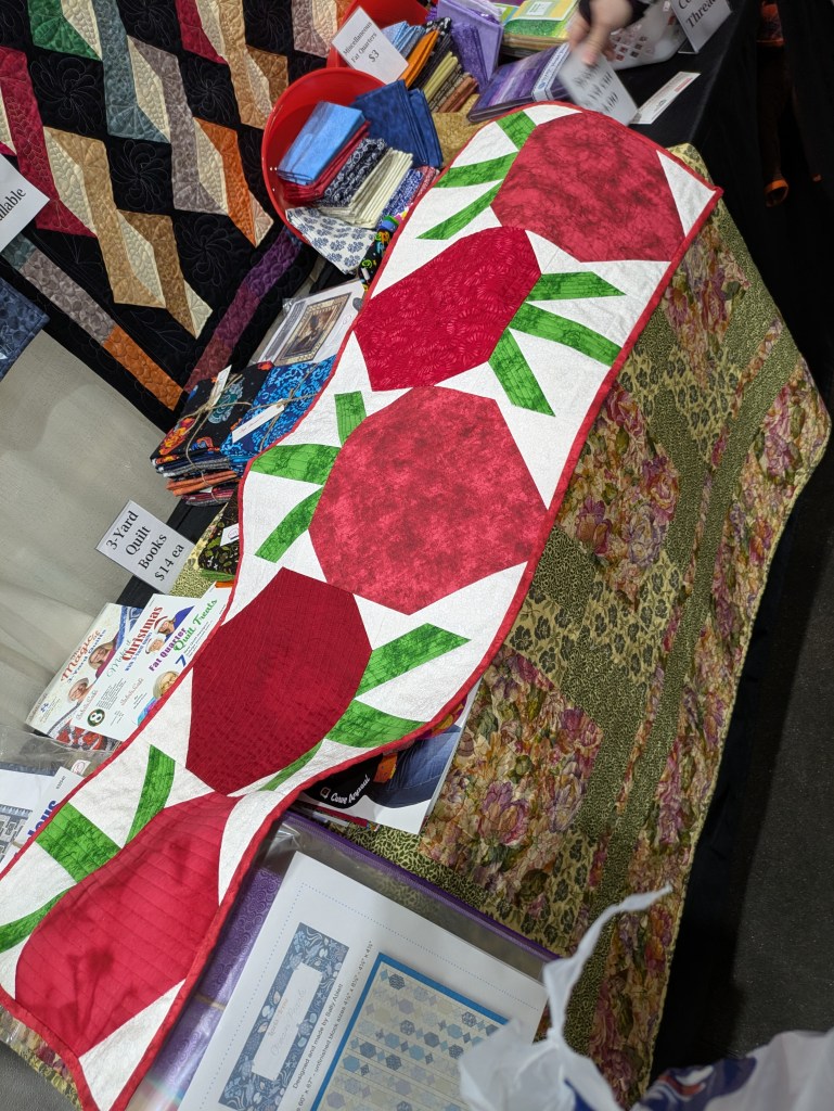 A colorful quilt display featuring a table runner with red round shapes and green leaves, alongside various fabric stacks in bright colors.