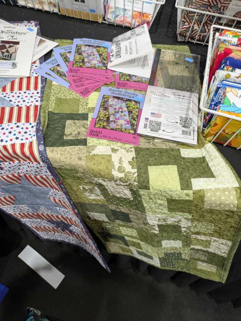 A green patchwork quilt displayed on a table, featuring various shades of green and intricate designs. Nearby, several quilting pattern booklets are spread out, showcasing quilt designs with colorful covers.