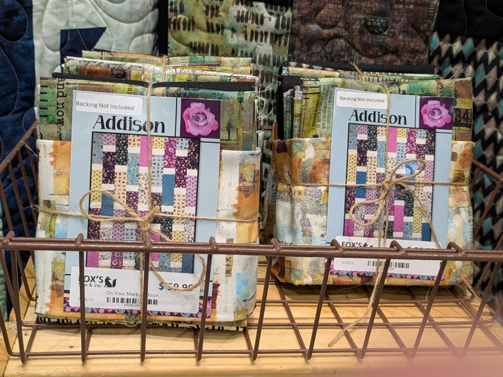 A basket containing two bundles of fabric squares tied with twine, labeled 'Addison'. Each bundle features colorful fabrics with different patterns, and a tag stating 'Backing Not Included'.