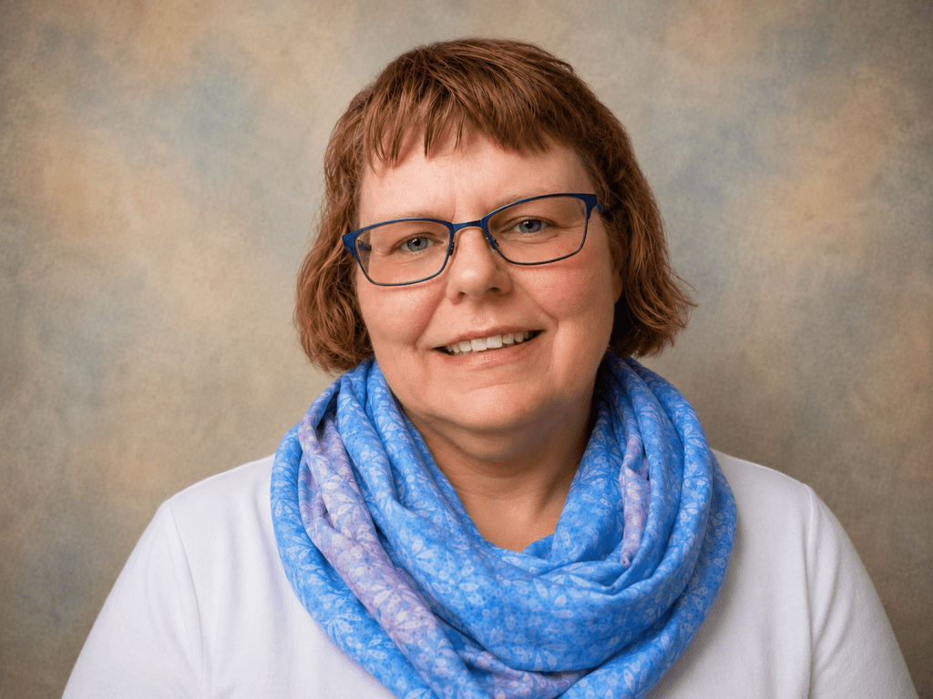 A smiling woman with short brown hair and glasses, wearing a blue and purple patterned scarf over a white top, posing in front of a softly blurred background.