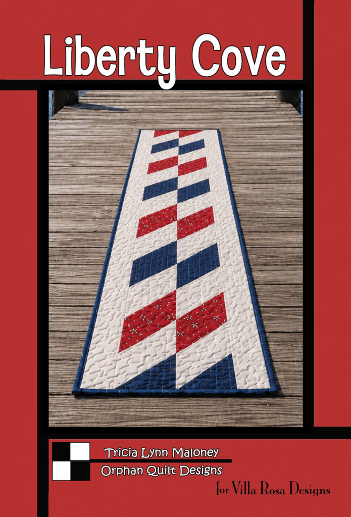 A quilted table runner featuring a geometric pattern in red, white, and blue colors, placed on a wooden surface, with text indicating the design name 'Liberty Cove' and designer details.