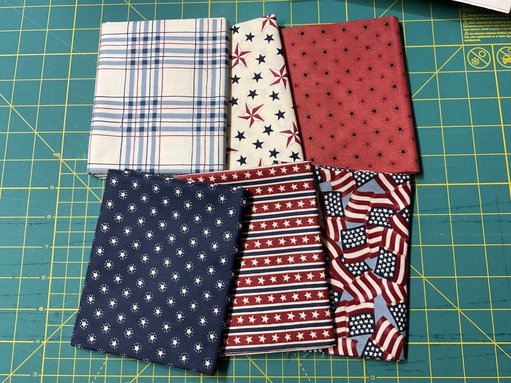 A collection of six fabric pieces featuring patriotic patterns, including plaid, stars, and American flags, arranged on a green cutting mat.