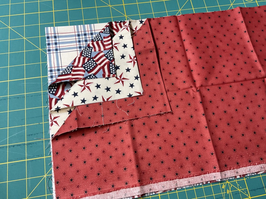 A piece of red fabric with a star pattern, layered with patterned fabric featuring American flags and stars, alongside a blue plaid fabric, on a green cutting mat with grid lines.