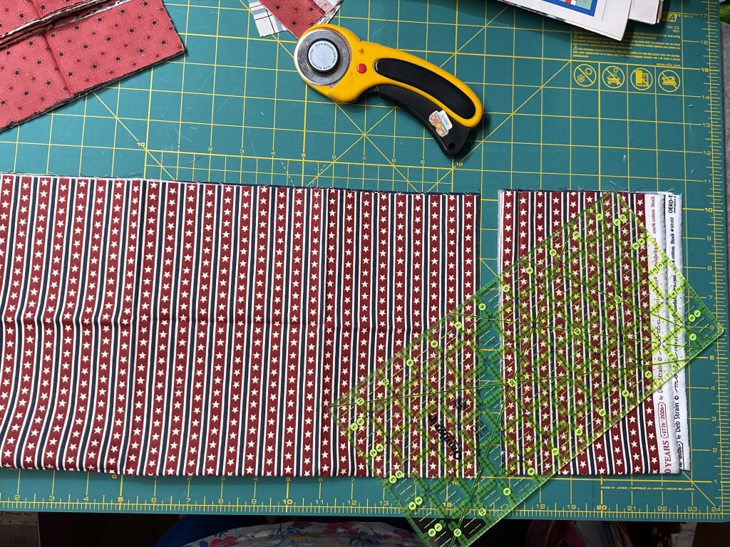 A green cutting mat with a rotary cutter, a patterned fabric piece in red and white stripes with stars, and a clear quilting ruler placed on the fabric.