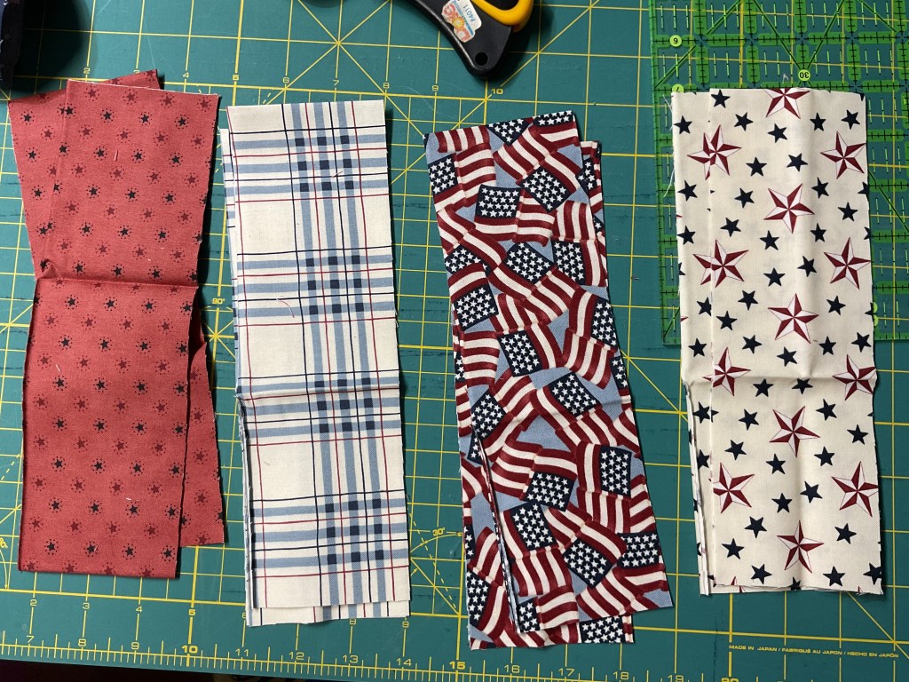 Four pieces of fabric in red, white, and blue patterns laid out on a cutting mat, featuring stars, stripes, and plaid designs.