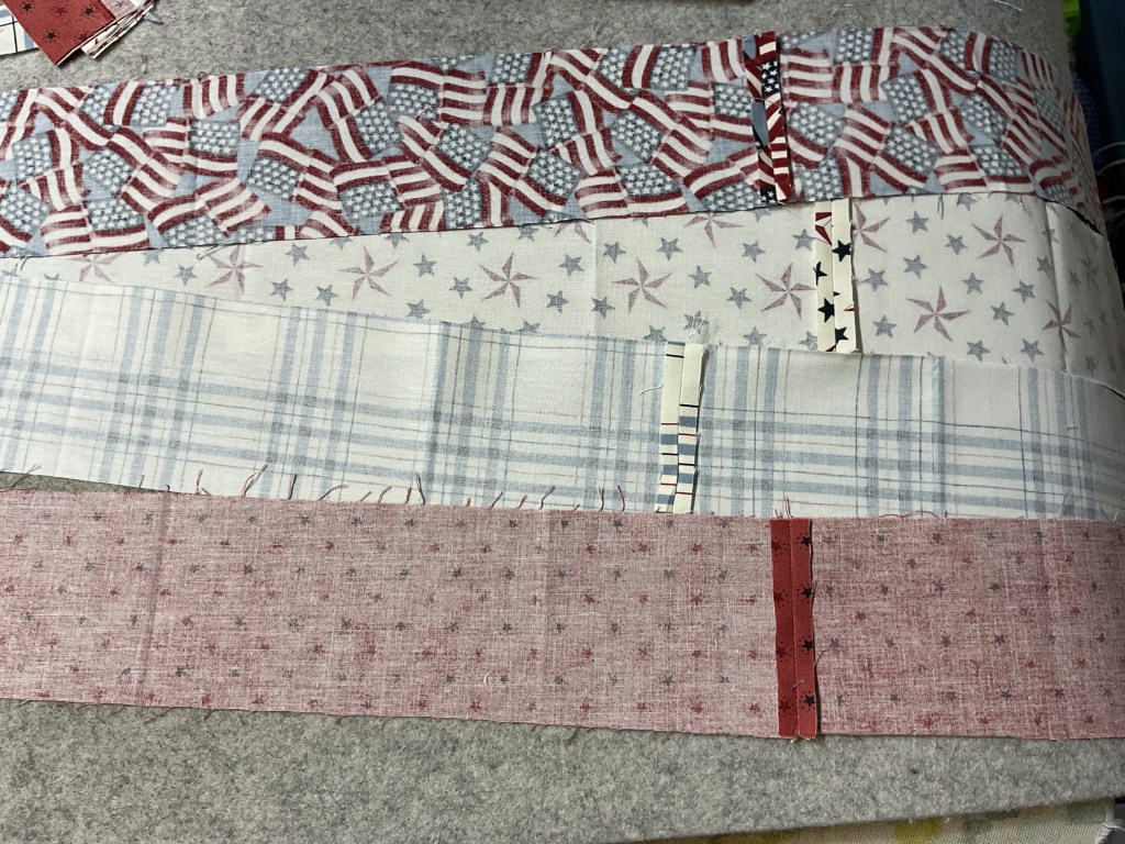 A collection of fabric strips featuring patriotic patterns, including American flags, stars, and plaid designs in red, white, and blue colors, arranged horizontally on a grey surface.