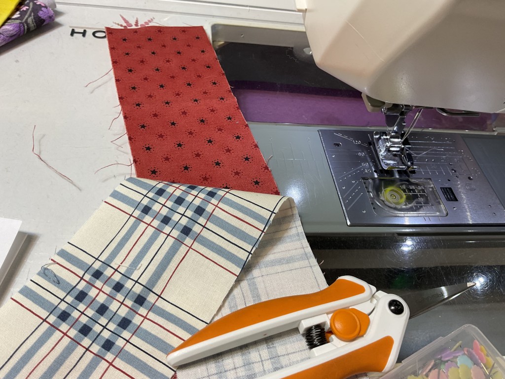 A sewing machine on a table with three different pieces of fabric, including a red patterned piece and a plaid piece, along with a pair of orange scissors.