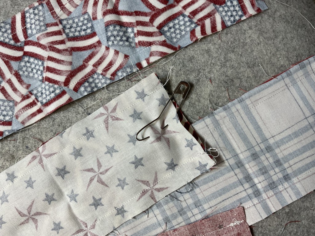 An assortment of fabric strips with patriotic patterns, including American flags and stars, pinned together with a safety pin, placed on a textured surface.