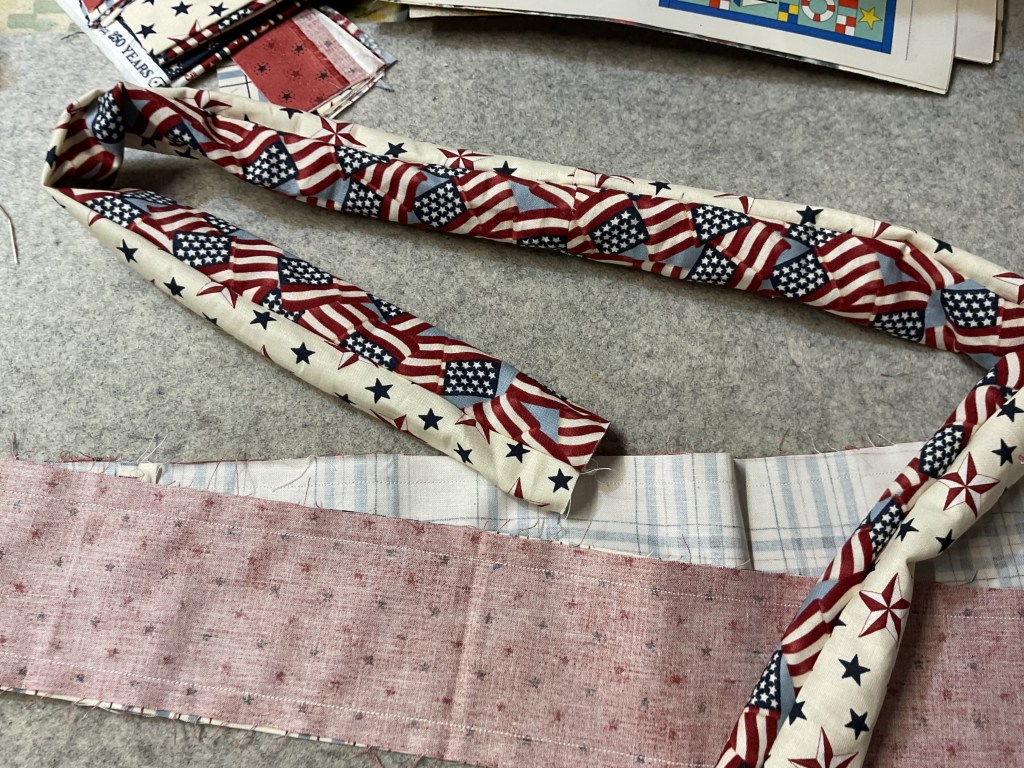 A close-up image of patterned fabric featuring American flags and stars, with additional fabric pieces in the background including plaid and star-patterned fabric.