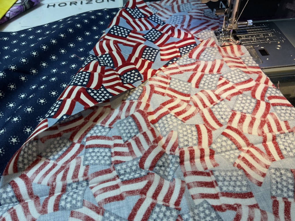 Close-up of fabric pieces featuring red, white, and blue patriotic patterns as well as quilting stars, alongside a sewing machine.