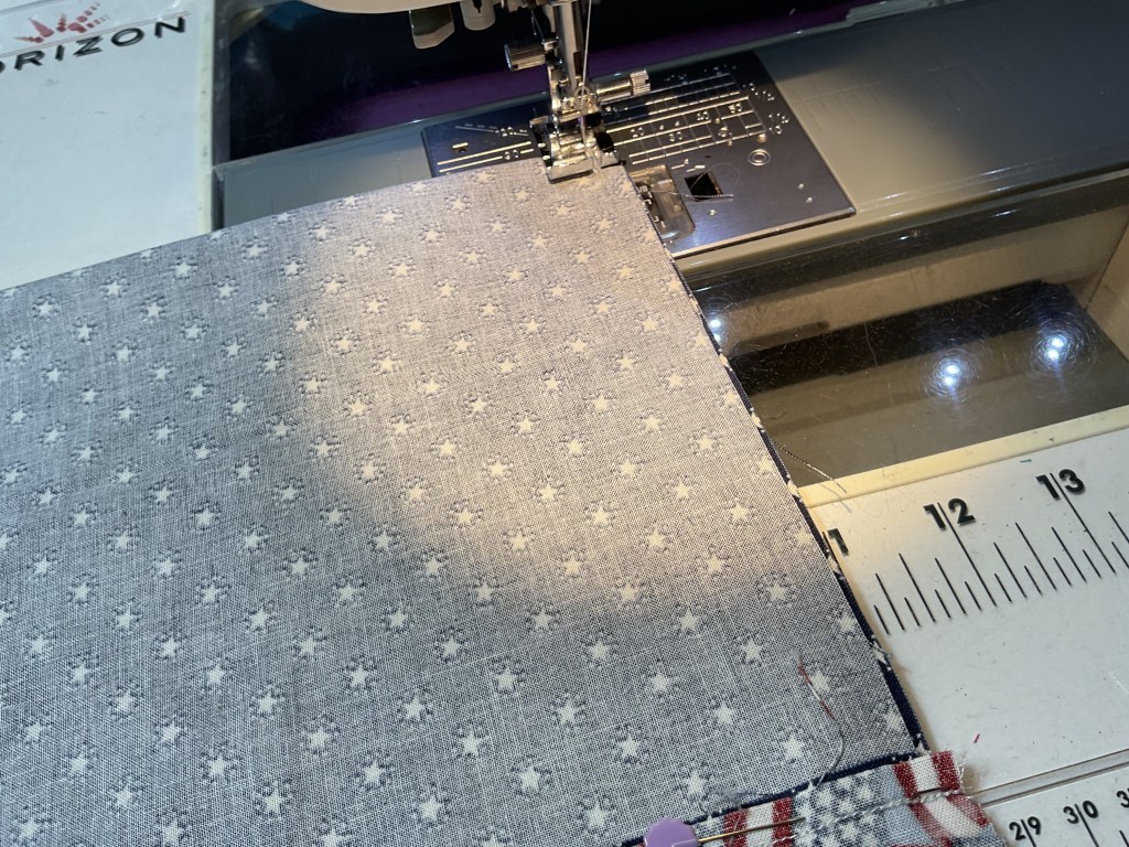 Close-up of a sewing machine with fabric under the needle, featuring a light gray patterned material. The machine is positioned on a work surface, with measuring lines visible in the foreground.