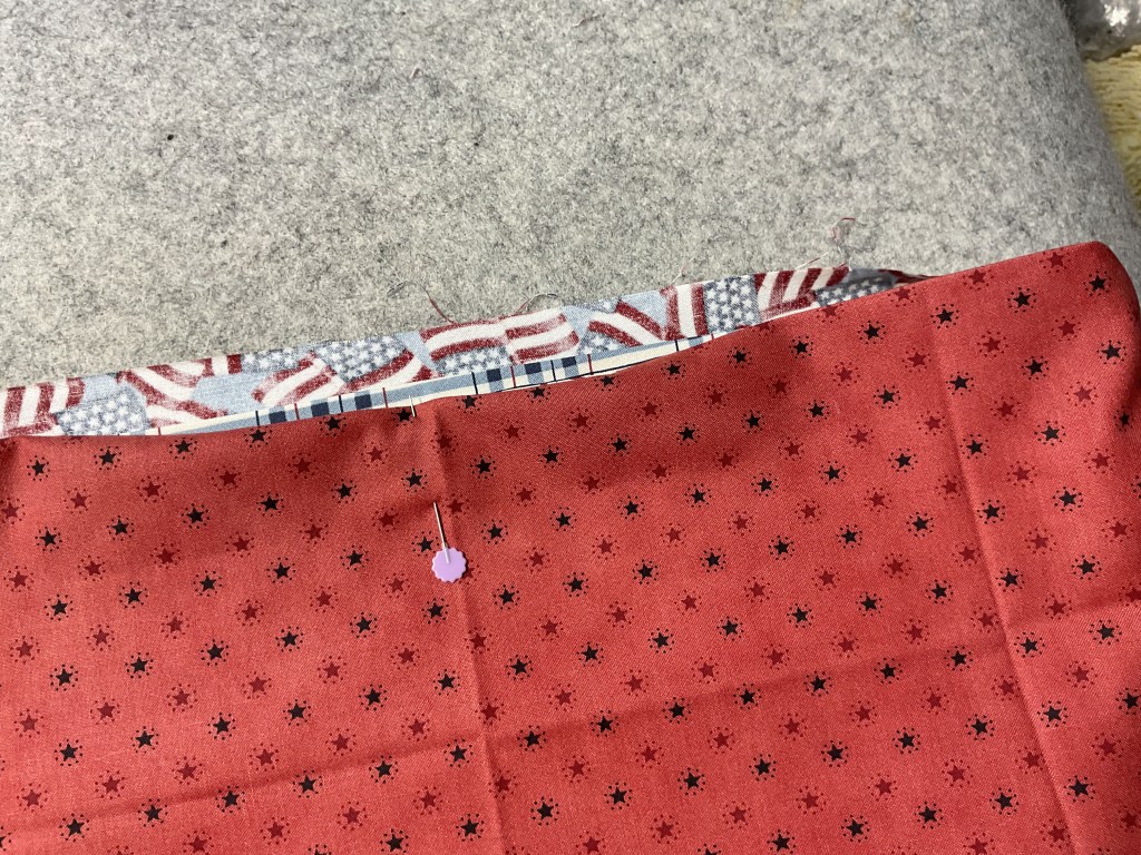 Close-up of a sewing project featuring red fabric with black star patterns, pinned to a blue fabric with red and white stripes, on a gray felt surface.