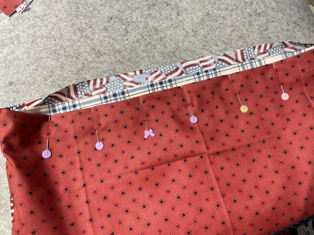 A piece of red fabric with a star pattern, pinned with colorful fabric clips for sewing, partially layered with a contrasting fabric featuring stripes and stars.