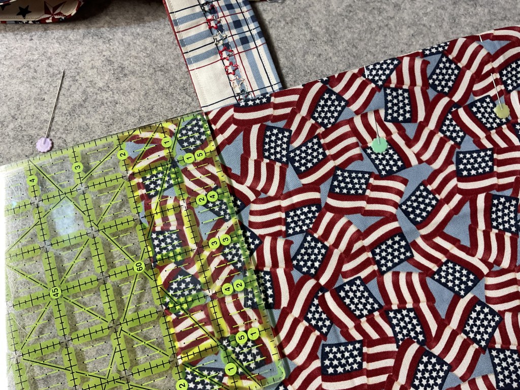 A close-up of a cutting mat with a ruler on top, featuring a patriotic fabric printed with American flags. A sewing needle and pins are also visible.