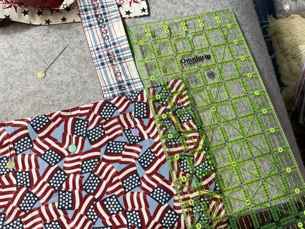 A close-up view of a quilting workspace featuring red, white, and blue fabric with American flag designs, a cutting mat with measurement grids, and pins holding the fabric in place.