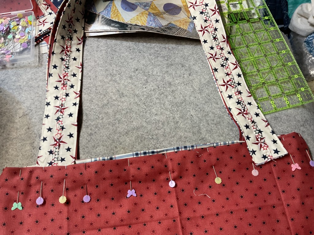 A close-up view of a partially assembled tote bag featuring a red fabric with stars, decorated straps with star patterns, and colorful pins securing the fabric pieces on a gray surface.