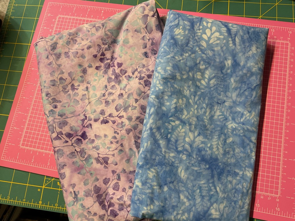 Two pieces of fabric on a cutting mat: one with a purple and lavender floral pattern, and the other in varying shades of blue with a leaf design.