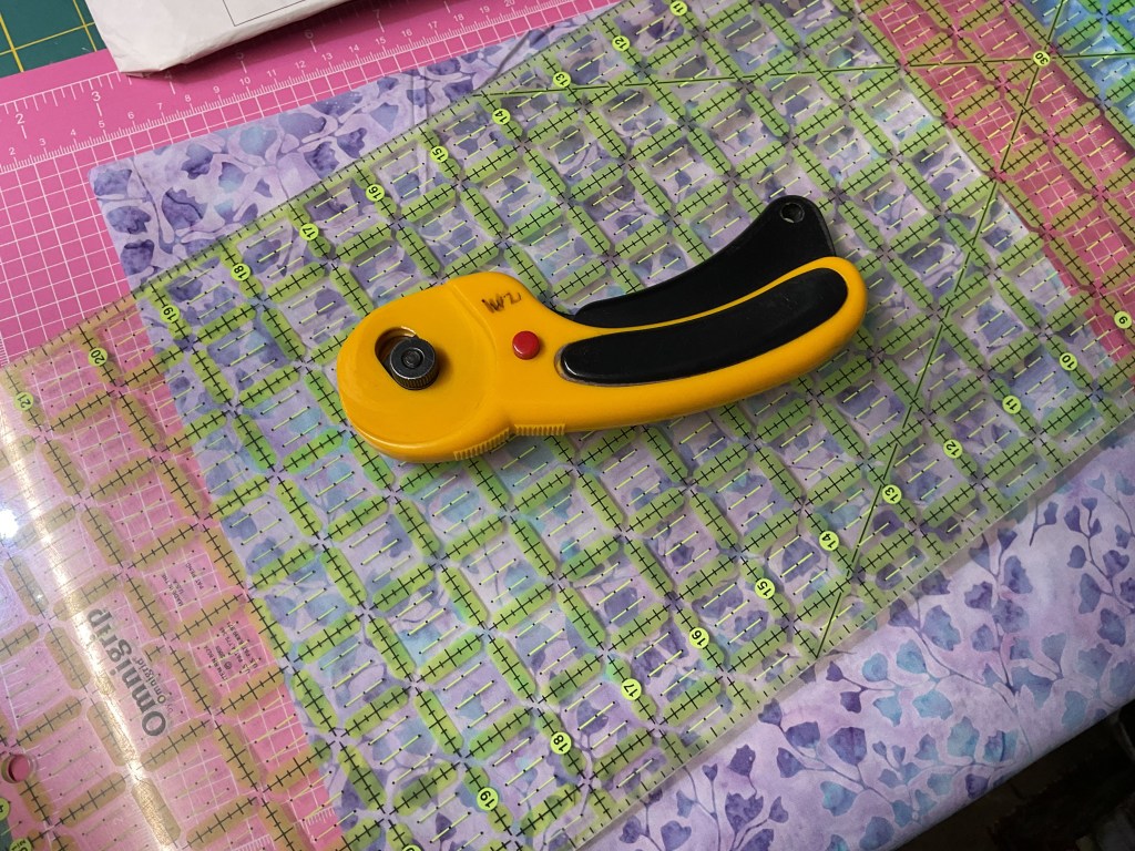 A rotary cutter placed on a cutting mat with a grid pattern, surrounded by layered fabric in various colors and patterns.