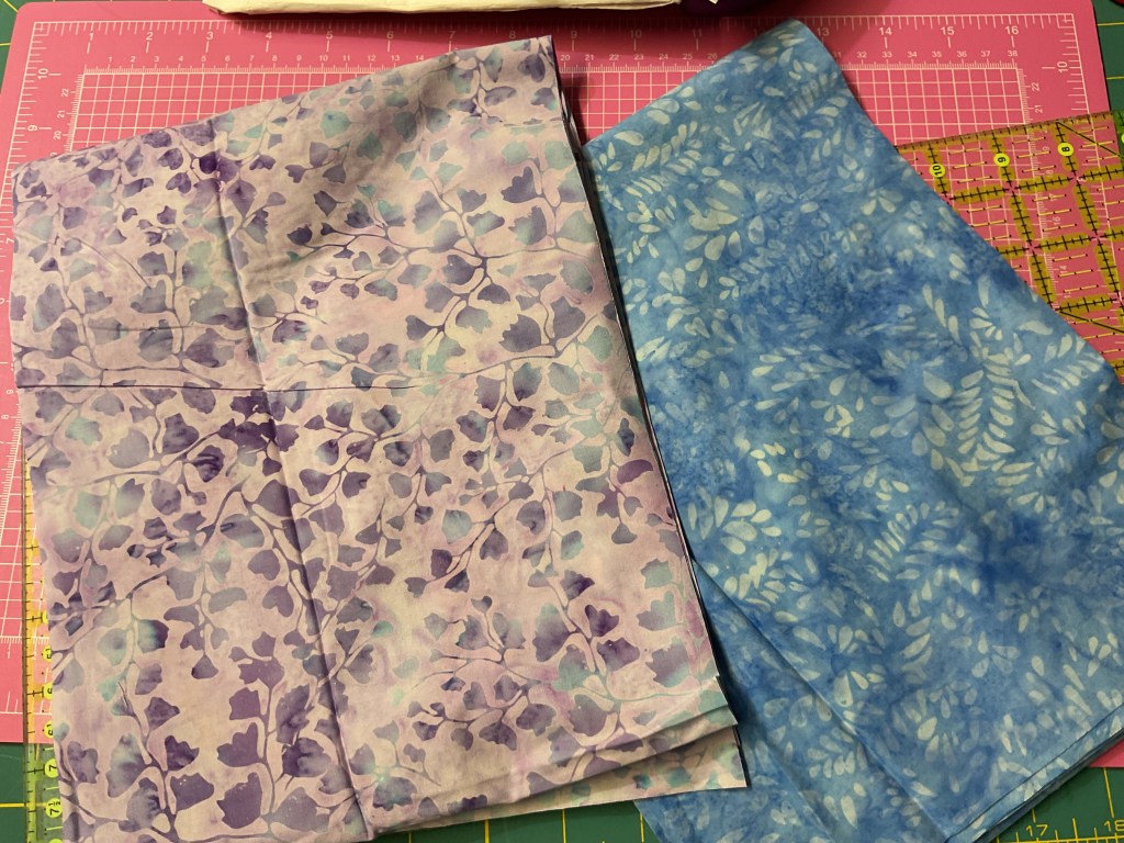 Two pieces of fabric laid out on a cutting mat; one has a floral pattern in purple and blue shades, and the other features a blue background with light leaf designs.
