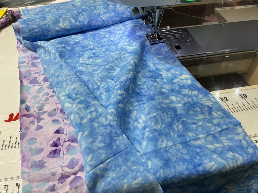 A close-up view of two pieces of fabric being sewn together on a sewing machine, featuring a blue patterned fabric and a purple floral fabric.