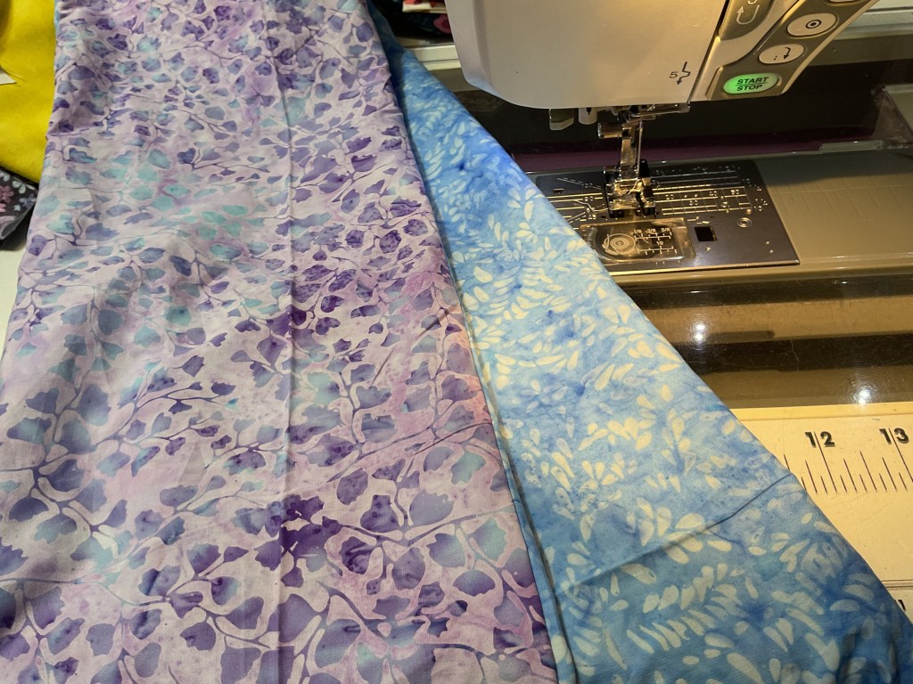 A close-up view of two pieces of fabric, one featuring a purple and blue floral pattern and the other in a blue tone with a subtle leaf design, placed next to a sewing machine.