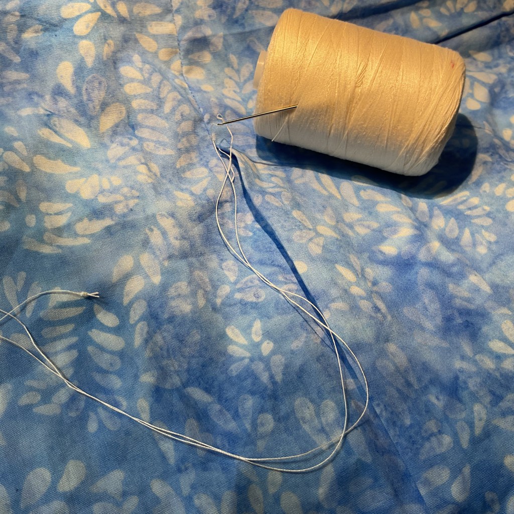 A sewing needle threaded with white thread lies on a blue fabric with a leaf pattern, alongside a spool of beige thread.