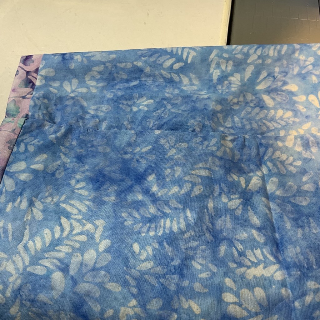 Close-up of a blue fabric with a leaf pattern, partially folded with a purple patterned edge visible.
