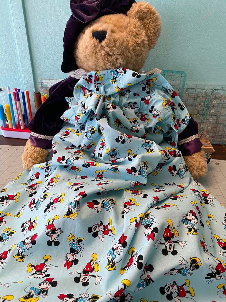 A teddy bear wearing a purple hat and dress made of light blue fabric featuring colorful Mickey and Minnie Mouse patterns.