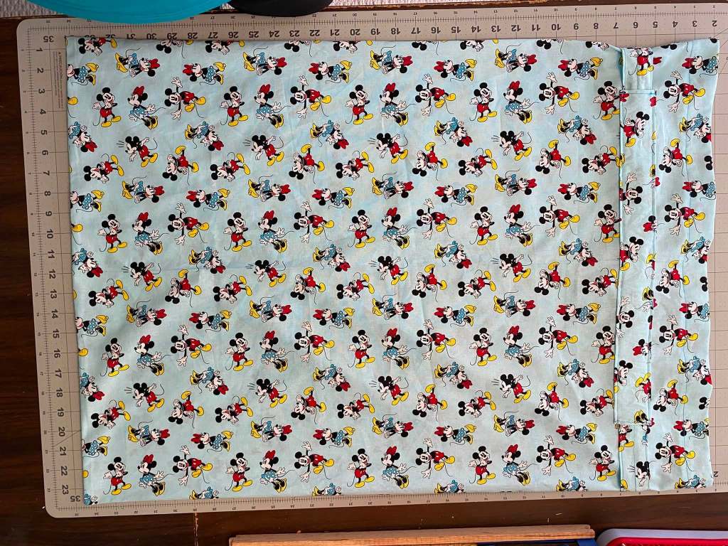 A piece of fabric featuring a repeating pattern of cartoon characters, including Mickey Mouse and Minnie Mouse, on a light blue background. The fabric is laid flat on a cutting mat with measurement markings.