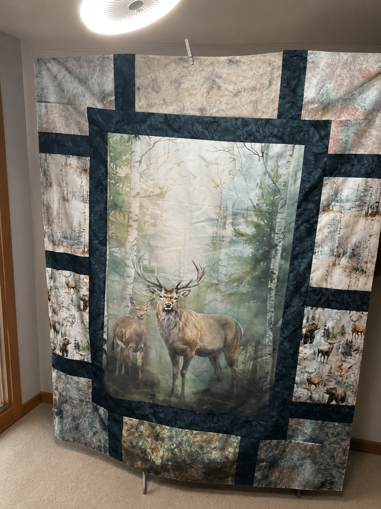A large quilt hanging on a wall, featuring a central image of two deer in a serene forest setting, surrounded by various nature-themed fabric panels depicting wildlife.