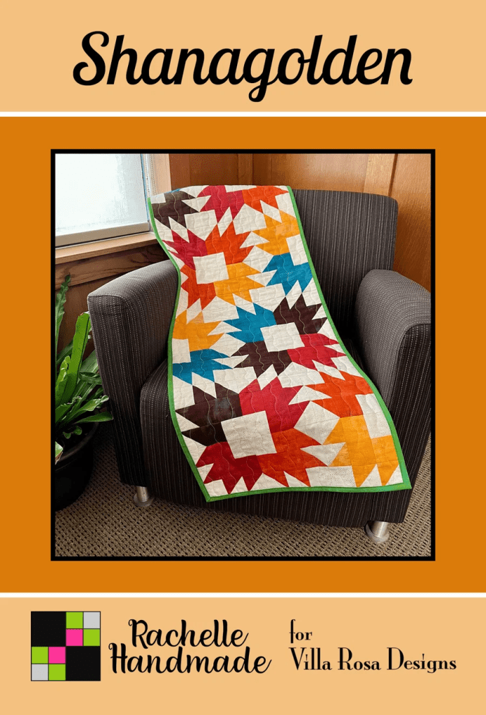 A colorful quilt with a star pattern, draped over a gray armchair, in a cozy indoor setting. The quilt features shades of red, orange, blue, and brown against a white background.