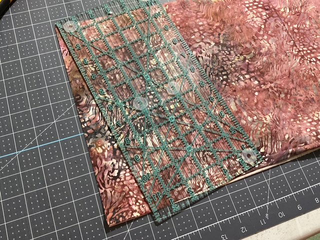 A transparent quilting ruler placed on patterned fabric with shades of pink, on a cutting mat with a grid layout.