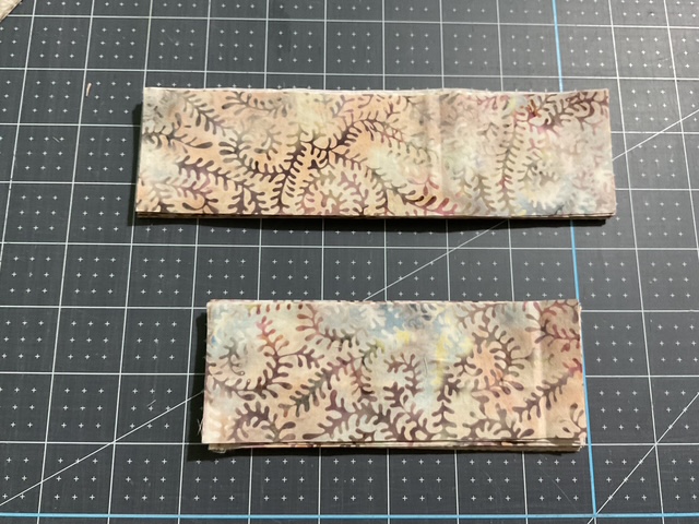 Two fabric pieces with a botanical pattern, one larger and one smaller, placed on a black grid cutting mat.