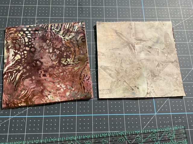 Two patterned squares of paper on a cutting mat. The left square features a vibrant mix of red, brown, and green abstract designs, while the right square has a softer, lighter pattern in neutral tones.