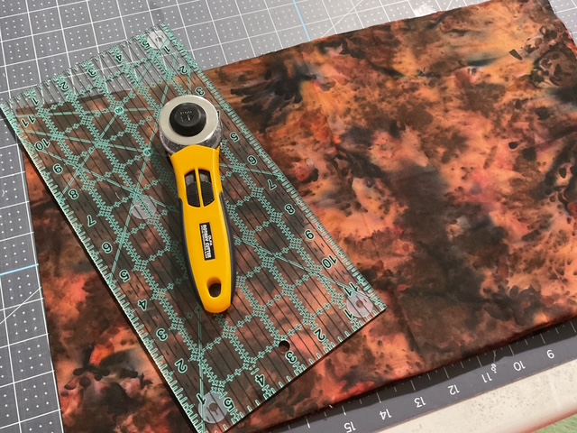 A rotary cutter and ruler resting on a piece of patterned fabric on a cutting mat.