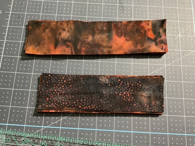 Two pieces of fabric displayed on a cutting mat; the top piece has a mottled pattern in shades of orange and brown, while the bottom piece features a black background with red dotted patterns.
