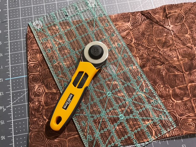 A rotary cutter and a transparent quilting ruler placed on a textured brown fabric.