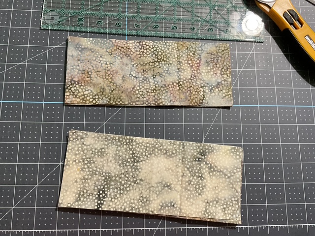 Two pieces of patterned fabric laid out on a cutting mat, with one piece displaying a light and dark speckled design and the other a lighter, more muted speckled pattern. A clear ruler and a utility knife are placed nearby.