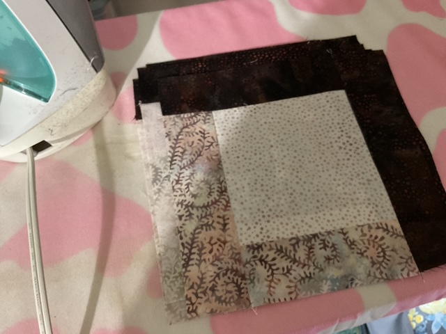 A stack of fabric squares in various colors and patterns, including a white square with gray dots and pieces with floral designs, placed near an iron on a pink and white patterned surface.