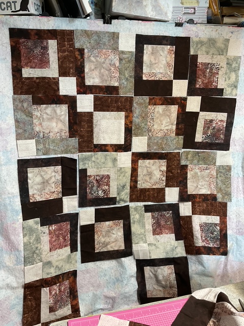 A patchwork quilt featuring a grid of squares in various shades of brown, green, and cream, with intricate patterns and textures.