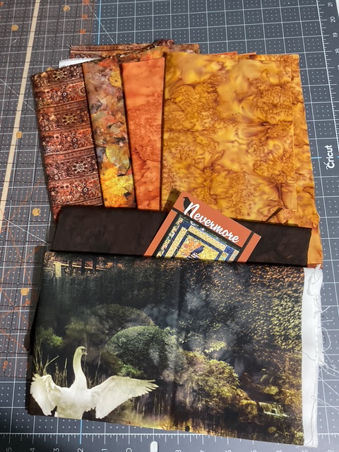 A collection of various fabric pieces in warm colors, including orange, brown, and patterned prints, arranged neatly on a cutting mat, with a small card labeled 'Nevermore' positioned among them.
