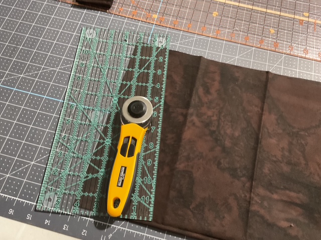 A rotary cutter and a transparent ruler positioned on a textured brown fabric, placed on a cutting mat with a grid pattern.