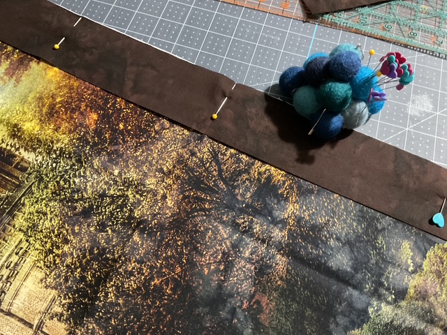 Close-up of a piece of fabric with a decorative autumn scene, pinned to a brown fabric at the top, with a colorful pom-pom pin holder nearby on a cutting mat.