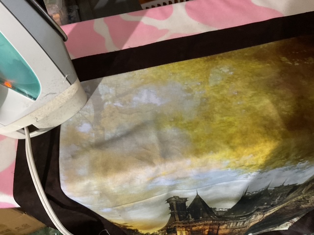 An iron resting on a colorful ironing board cover featuring a scenic painting with a structure and a sunset background.