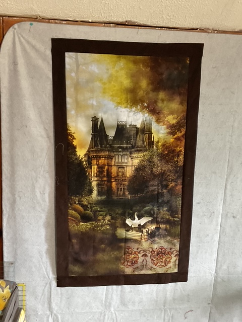 A decorative wall hanging featuring a picturesque castle surrounded by lush trees and a serene lake with two swans.