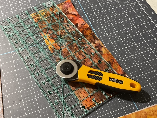 A rotary cutter and a transparent ruler positioned over a piece of fabric, set on a cutting mat with a grid pattern.