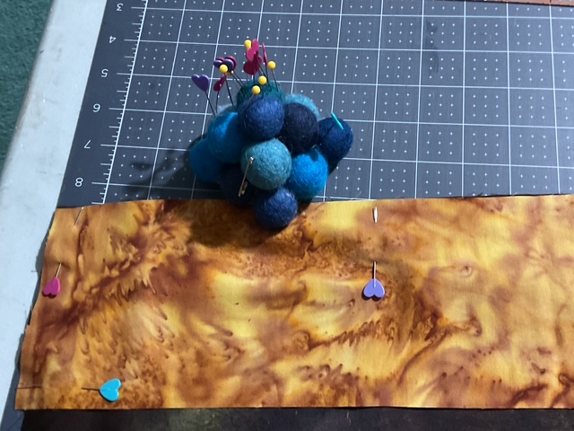 A sewing workspace showing a piece of fabric with a marbled brown pattern, pinned with colorful pins, next to a cluster of felt balls in various shades of blue.