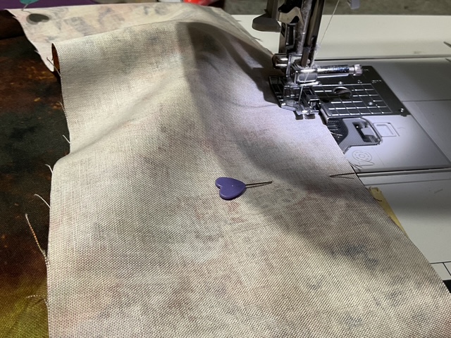 Close-up of a sewing machine needle positioned over a piece of fabric with a purple heart-shaped pin placed nearby.