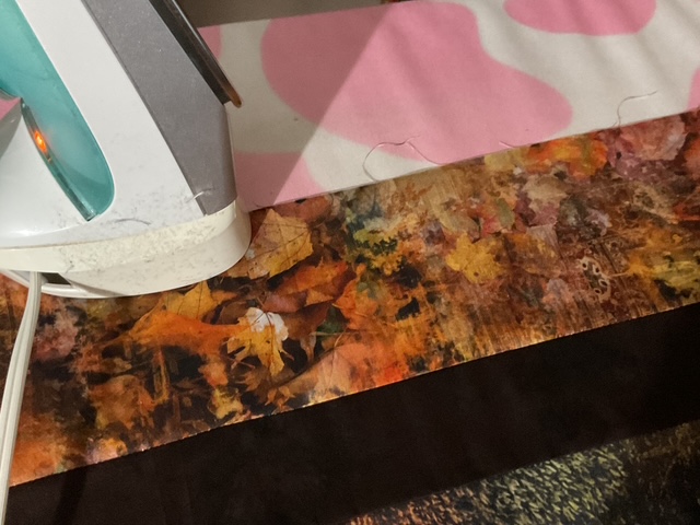 A close-up of an iron on a multi-colored fabric with a pattern of fall leaves, resting on a pink and white abstract patterned cloth.